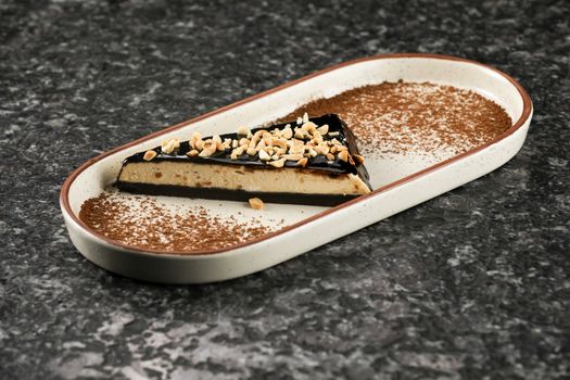 Entremets géométrique au chocolat-café, pécan, servi sur une assiette avec des éclats de noix et de la poudre de cacao.