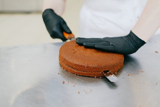 Un pâtissier en gants noirs découpe un gâteau en forme géométrique sur une surface en inox.