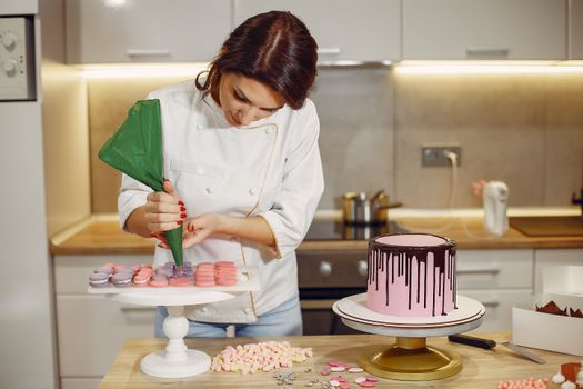 Une pâtissière prépare des macarons colorés et un gâteau rose avec un glaçage au chocolat dans une cuisine moderne.